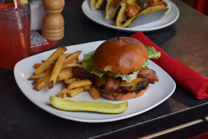 A close up of a bacon cheeseburger with fries and a pickle on the side.