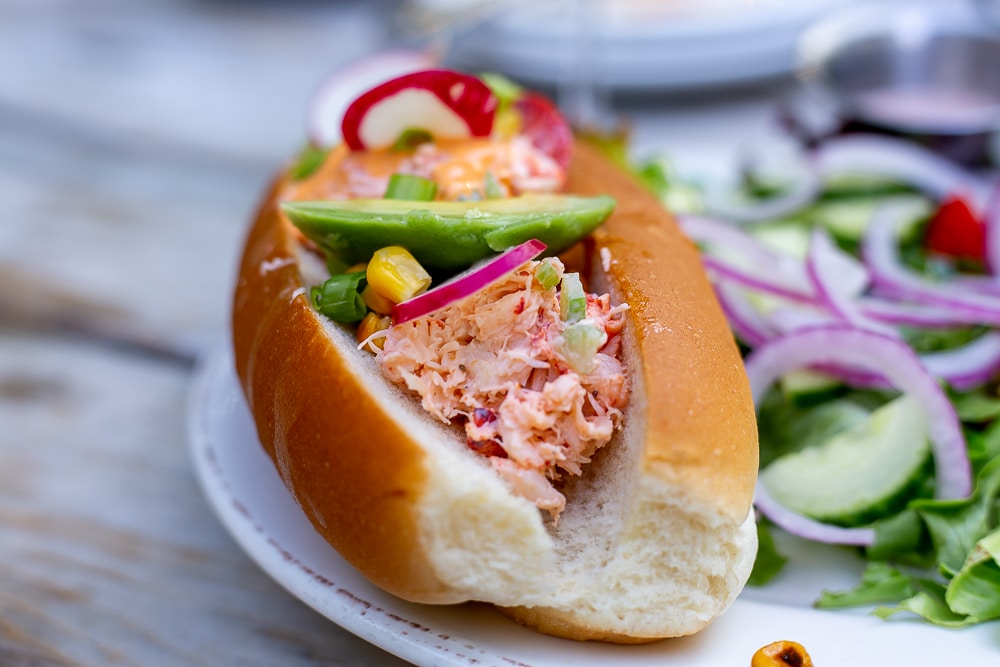 Lobster roll topped with avocado, corn, chive and summer radish next to a colorful side salad