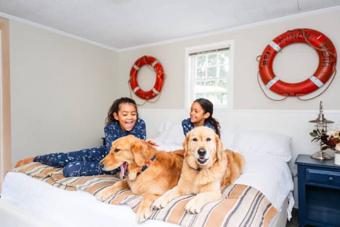 Sisters sitting on the edge of a queen bed in matching blue pajamas laughing while petting two golden retriever dogs laying at the foot of the bed.