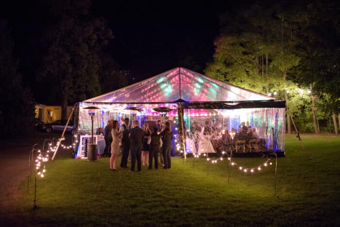 A wedding reception happening under a clear wedding tent with pink and green lights.