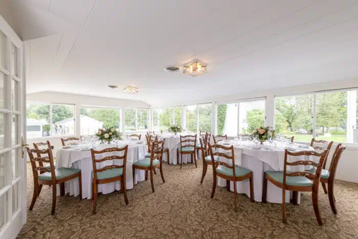 Several round tables with white table clothes set up for an event with flower centerpieces. The tables are in a room with large square window all around that have a view of a lawn.
