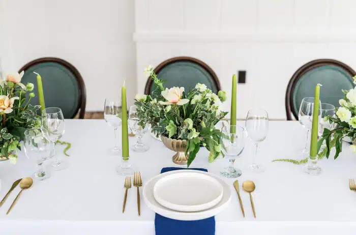 A close up of the flower arrangement and centerpieces on a rectangular table set for an event. The flower arrangements run down the middle of the table and have a long green candle set on either side of each flower arrangement.
