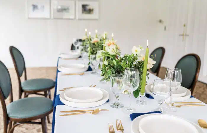 A close up of a rectangular table set up for an event with pink and white flower centerpieces running down the middle with long green candles on wither side of the flower arrangements.