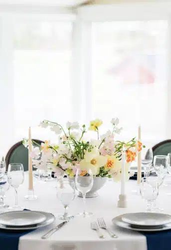 A close up of a round table with a yellow and pink flower centerpiece set up for a wedding.