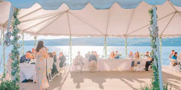 A woman giving a speech at a wedding with the guests watching under a wedding tent next to the lake.