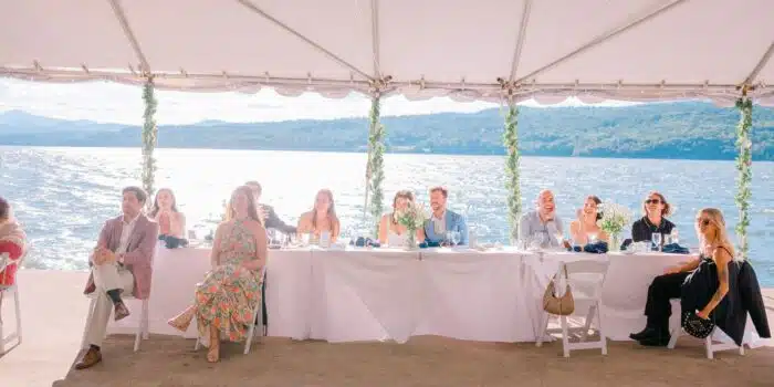 A wedding group sitting at a table laughing with a lake and mountains in the background.
